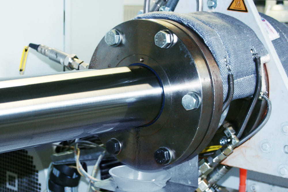 Close-up of industrial machinery showing a shiny metal piston connected to a large round flange with bolts. The setting is technical and precise.