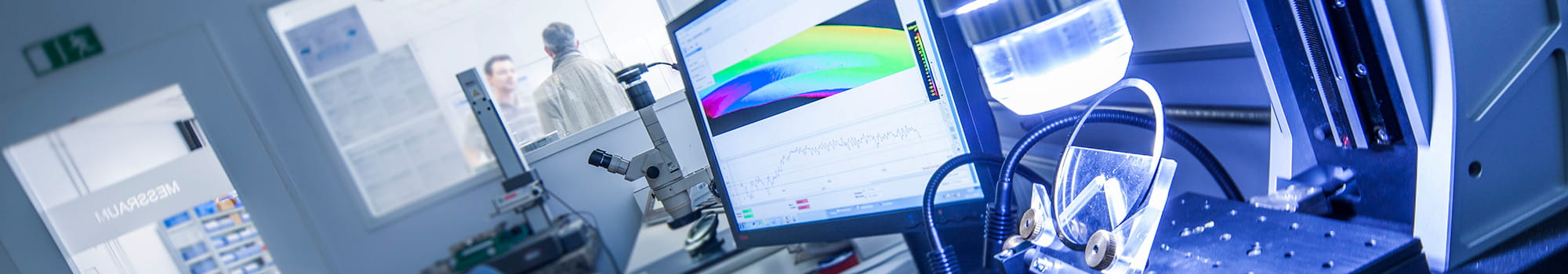A close-up of a high-tech laboratory setup with precision instruments, a computer displaying colorful data curves, and two people discussing in the background behind a glass wall.